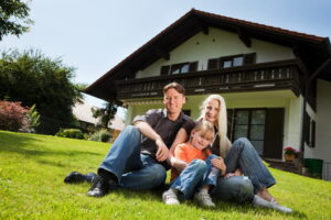 Happy family of three sitting on a well-manicured lawn in front of a modern two-story house with wooden balcony and large windows.
